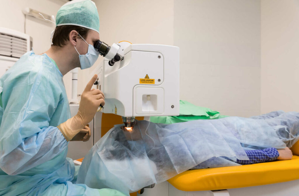 A male doctor in a blue and green surgical gown is sitting and his eyes resting on the eyepiece of a laser surgical machine while performing laser eye surgery on his patient.