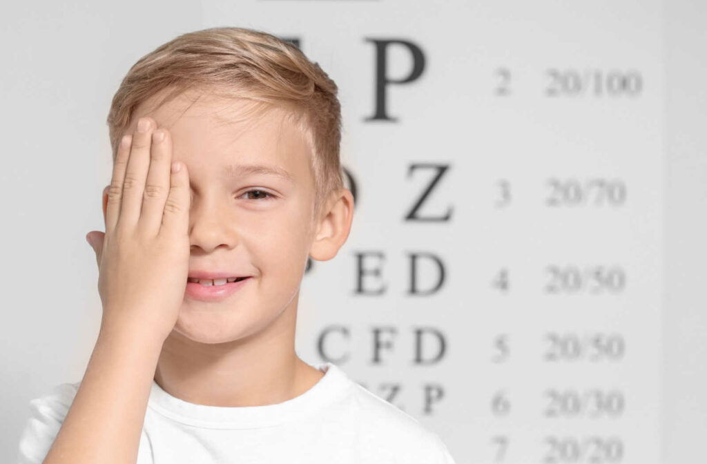 Smiling young boy standing in front of an eye chart and covering his right eye with his right hand.