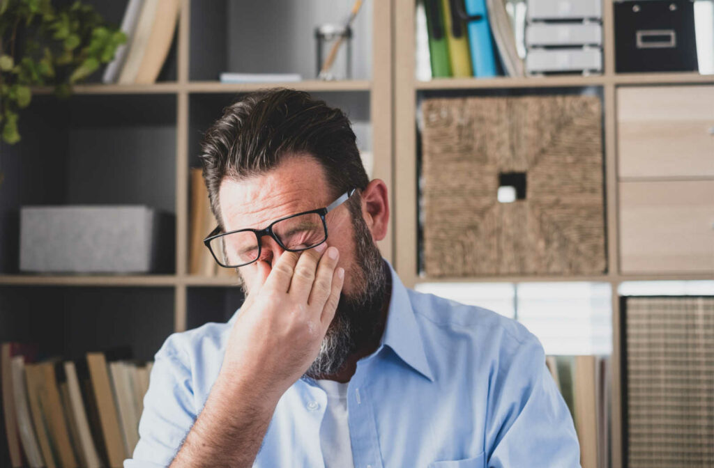 A man inside an office pushing his glasses up to rub his eyes with his fingers.