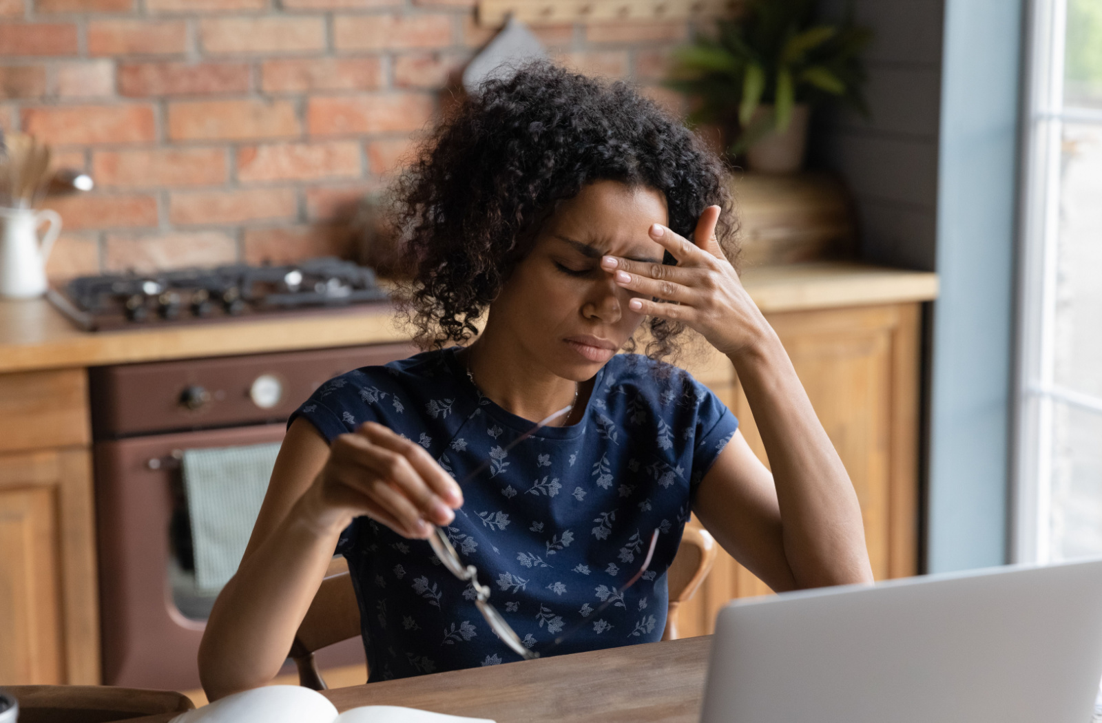 A woman sitting at a table in front of a laptop, holding her glasses in her right hand while pressing her left hand to her forehead as if in pain.