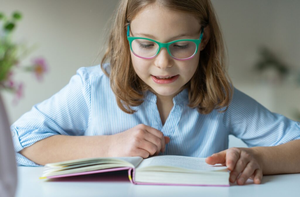 A young child with green and pink reading glasses sits at a desk reading a book with a pink cover.