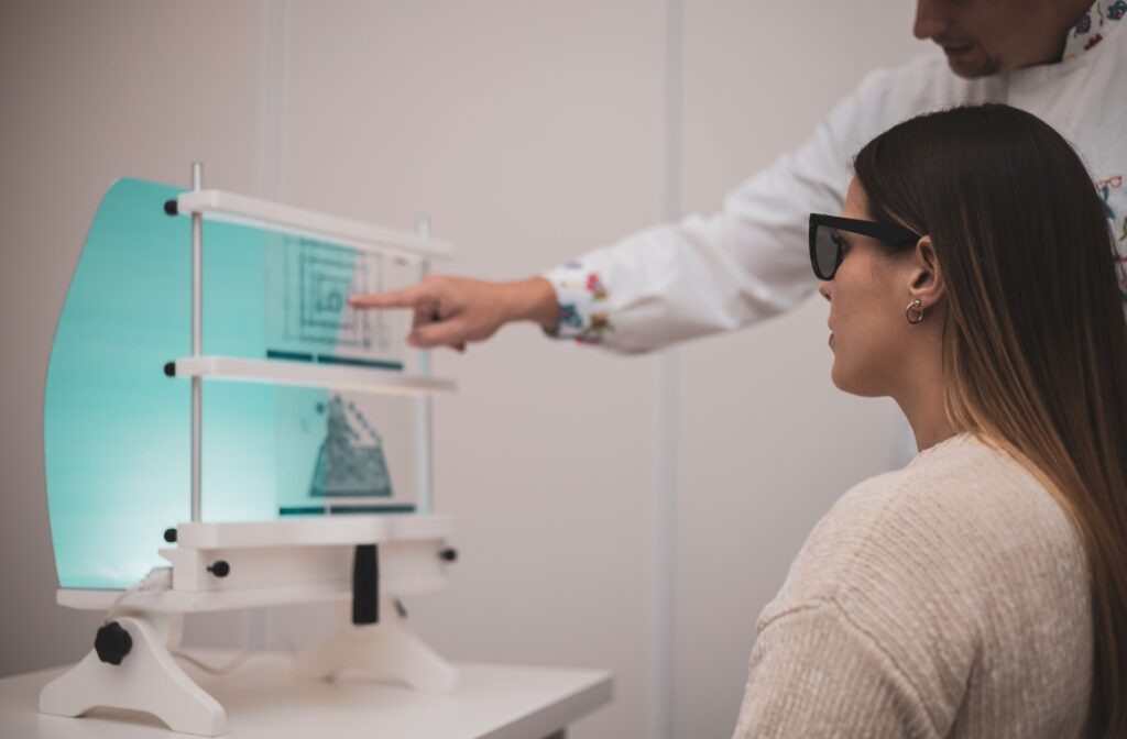 A woman wearing dark glasses sits in front of a stereoscopic vision testing device while a medical professional in a white coat points to a chart on the device. The setup is used for assessing depth perception and binocular vision.
