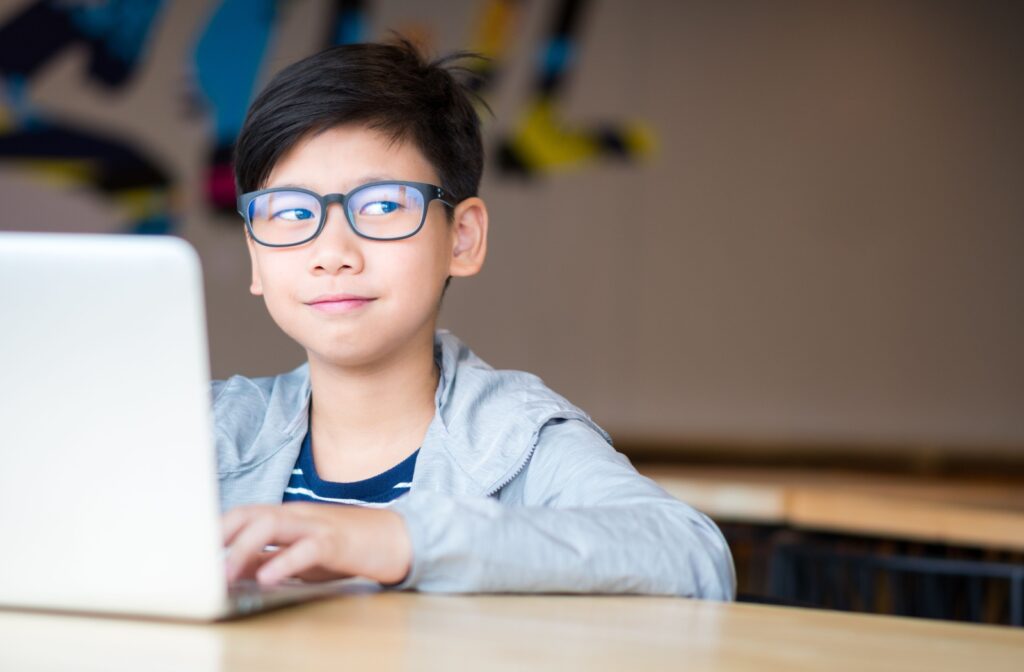 School-aged child with glasses typing on a laptop with a slight smirk.