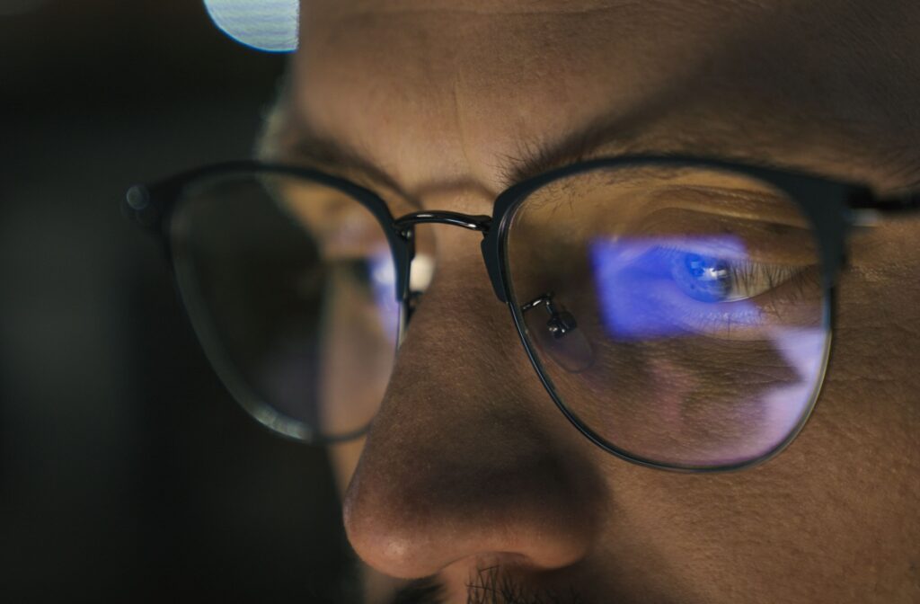 Close-up of a man wearing glasses with blue light from a computer screen reflecting on the lenses.