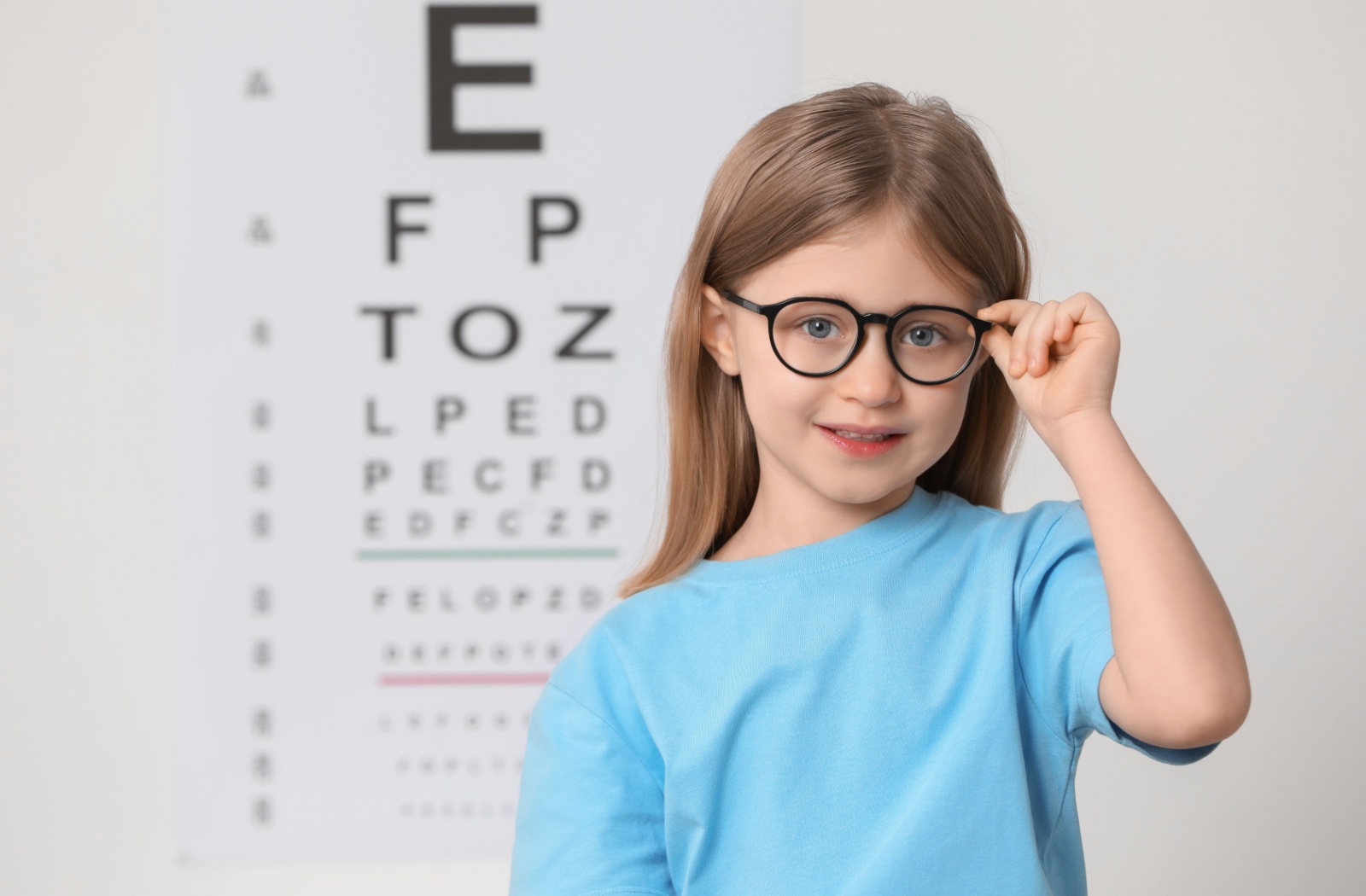 Young girl wearing glasses and smiling during an eye exam with a Snellen eye chart in the background.