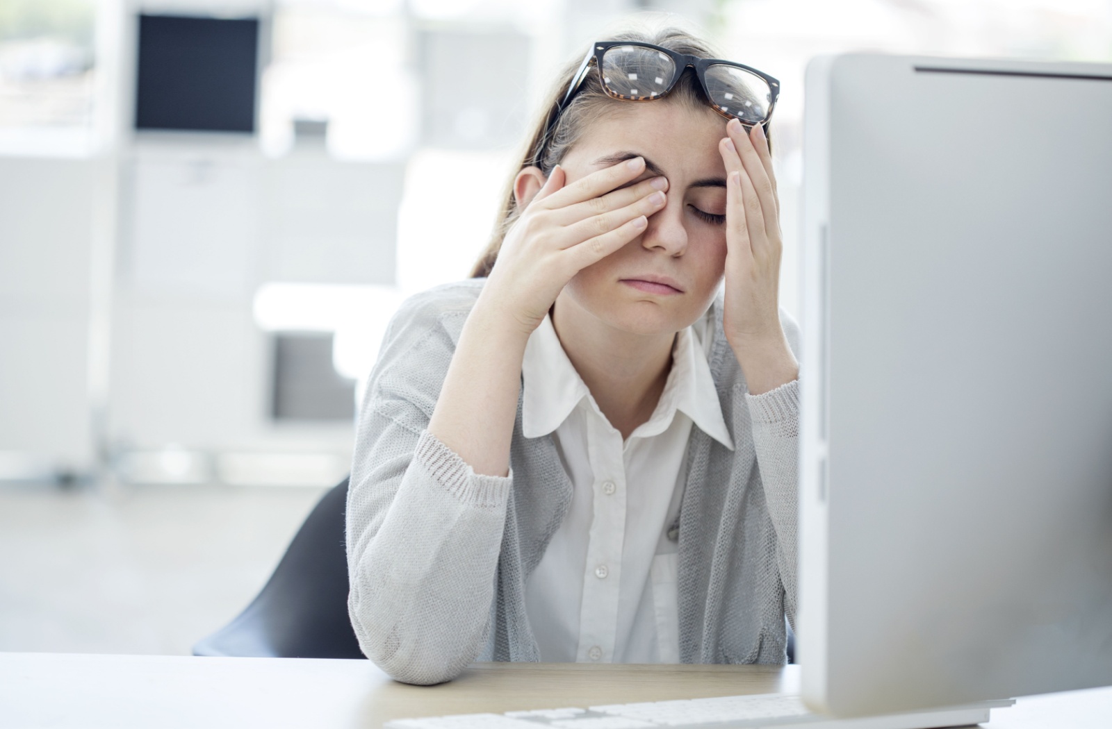 Woman sitting at a desk rubbing her eyes in front of a computer, showing signs of digital eye strain.