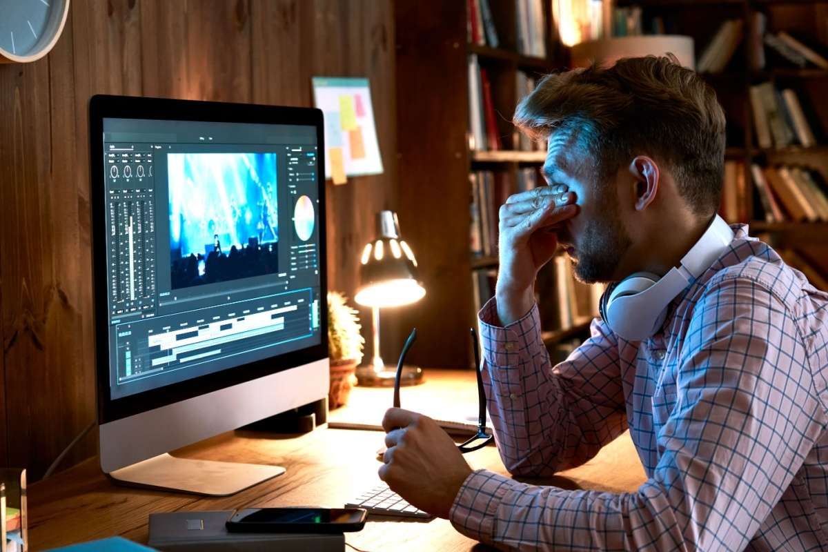 A man sitting at a desk rubbing his eyes while working on a large computer screen, showing signs of digital eye strain.