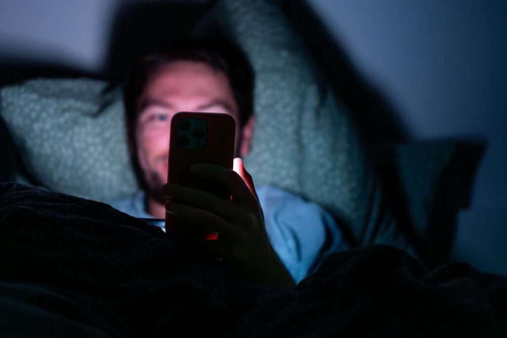 A man lying in bed using a smartphone in a dark room, with the screen lighting up his face and contributing to nighttime blue light exposure.