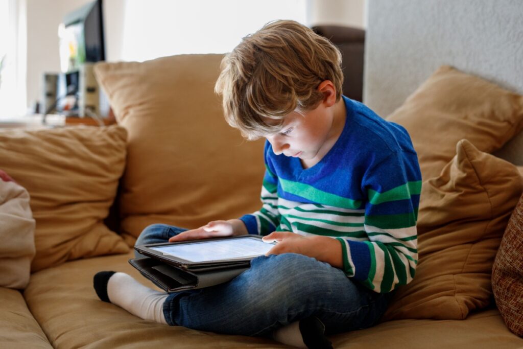 Young boy in striped shirt using tablet device while sitting cross-legged on couch at home.
