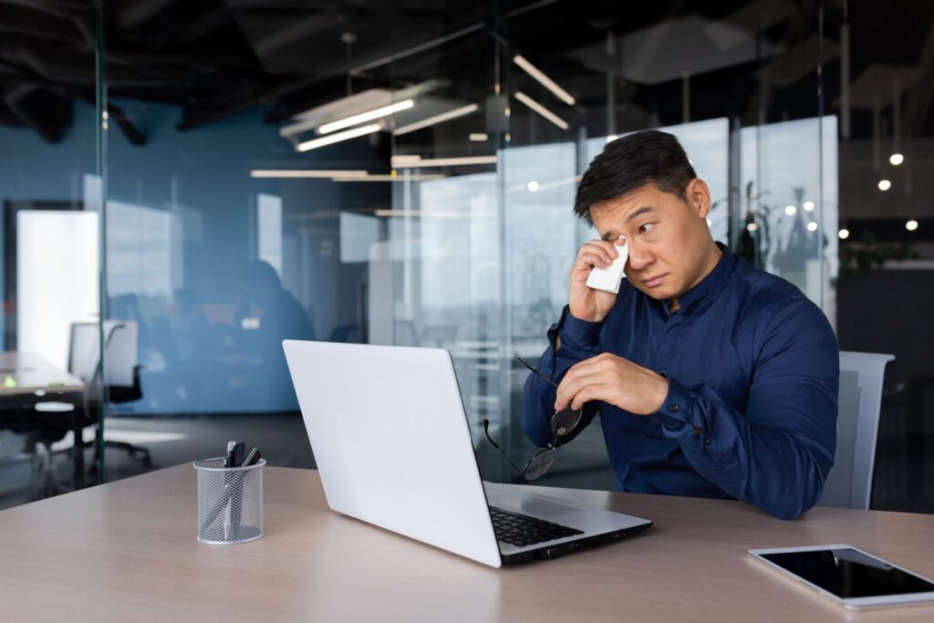 A man wiping his right eye while working at laptop computer in modern office environment.