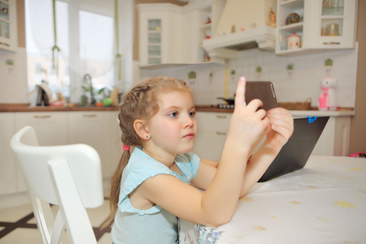 Young girl with braided hair holding tablet device while sitting at kitchen table in bright modern home.