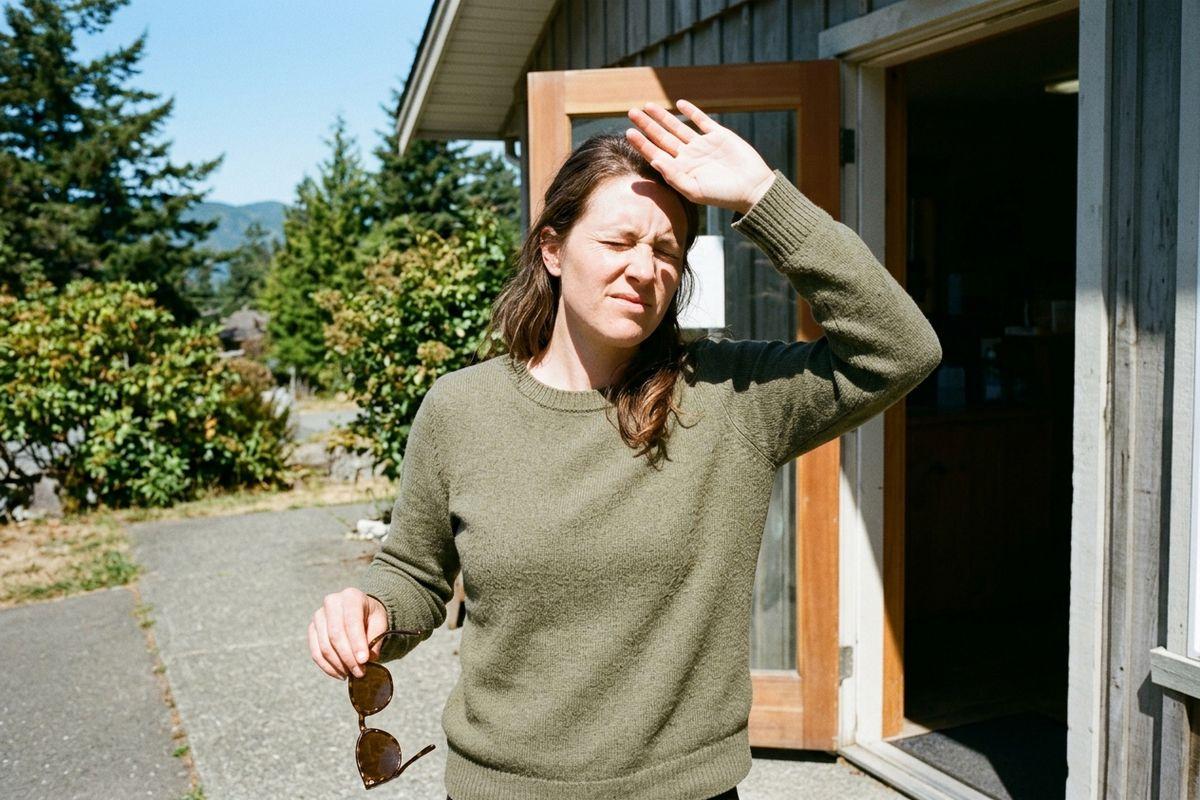 A person in a green sweater squinting outside a house doorway, shielding eyes with one hand and holding sunglasses in the other.